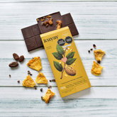 Kuyay Schokolade mit Ananas, 70% Kakao, auf einem hellen Holzuntergrund. Die Schokoladentafel ist geöffnet und zeigt Ananasstücke in der Schokolade, umgeben von getrockneten Ananasstücken und rohen Kakaobohnen als Dekoration.